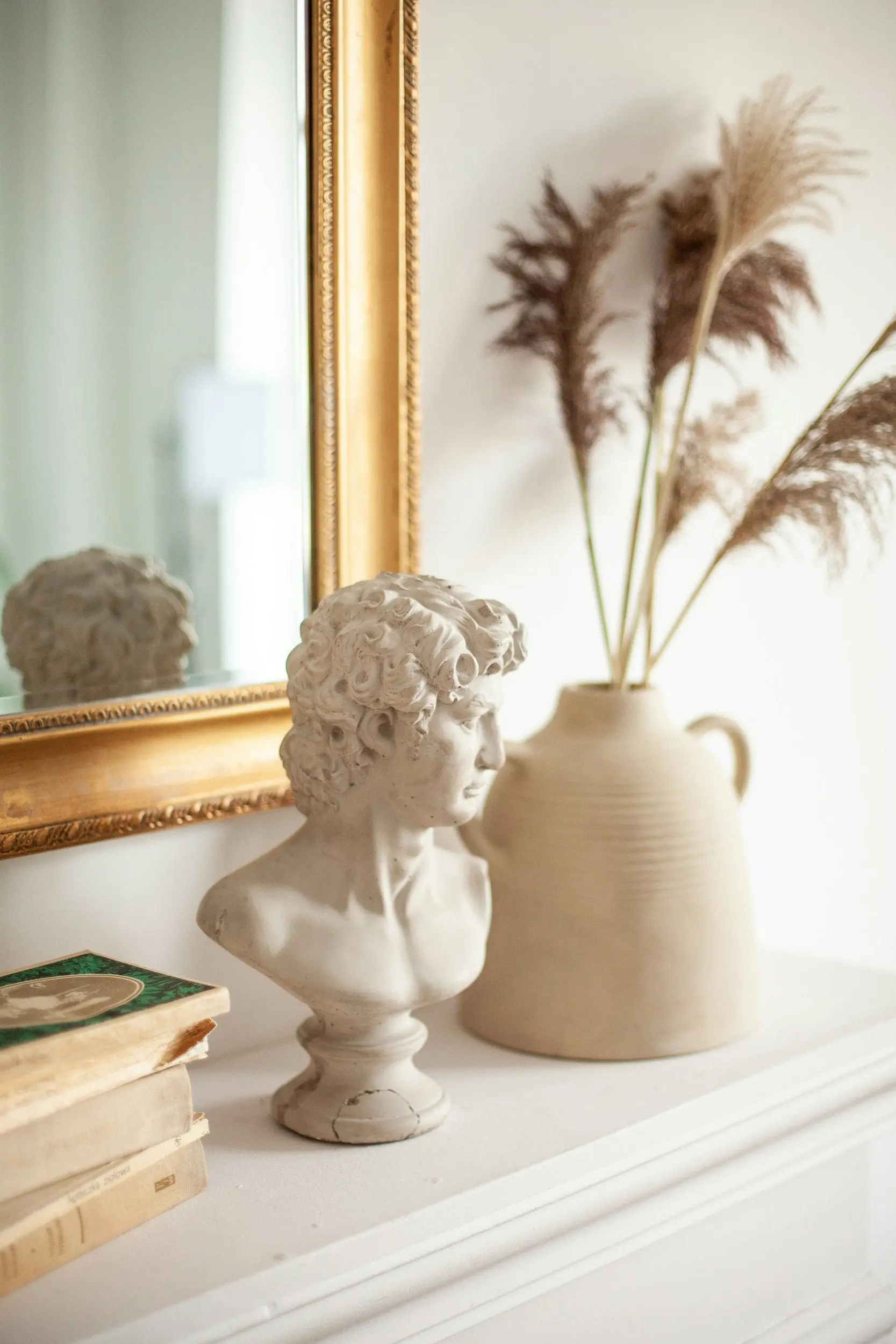 Classic white bust sculpture styled on a mantel with gold framed mirror, pampas grass in ceramic vase, and vintage books